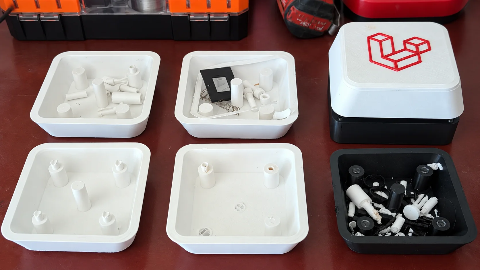 Several open 3D-printed button enclosures containing failed pins, springs, collars, screws, and switch parts beside the finished button